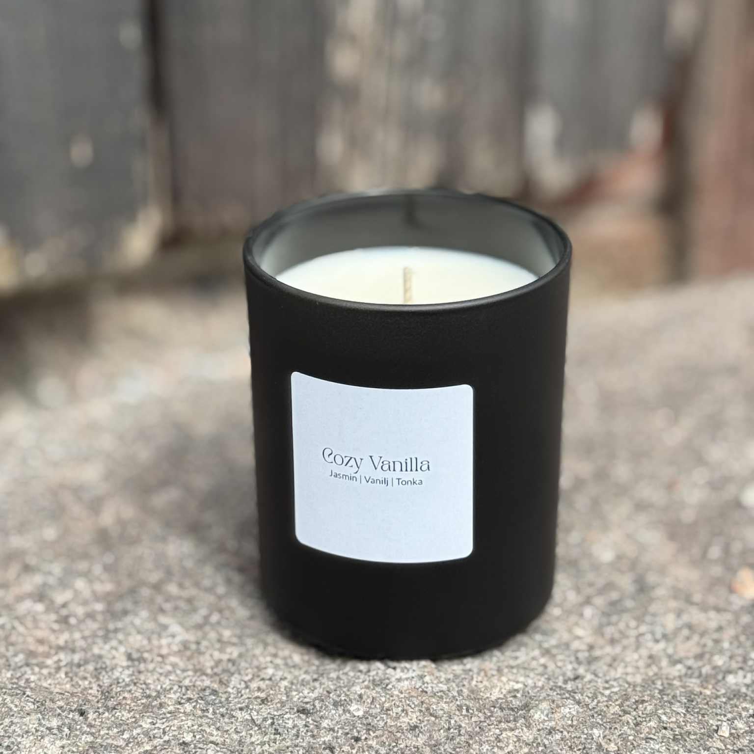 Doftljuset Cozy Vanilla i mattsvart glas placerat på sten med väderbitet trä i bakgrunden.