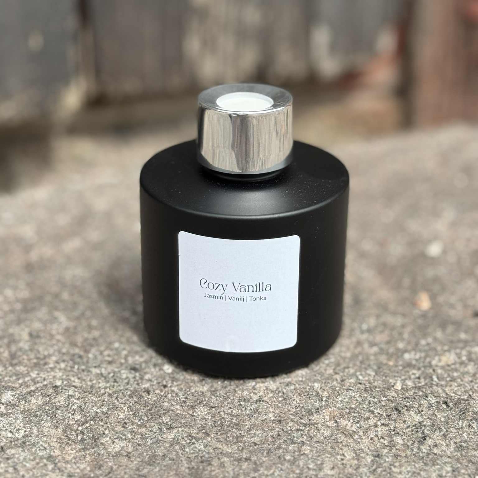 Cozy Vanilla doftpinnar i mattsvart flaska med silvertopp mot rustik träbakgrund.