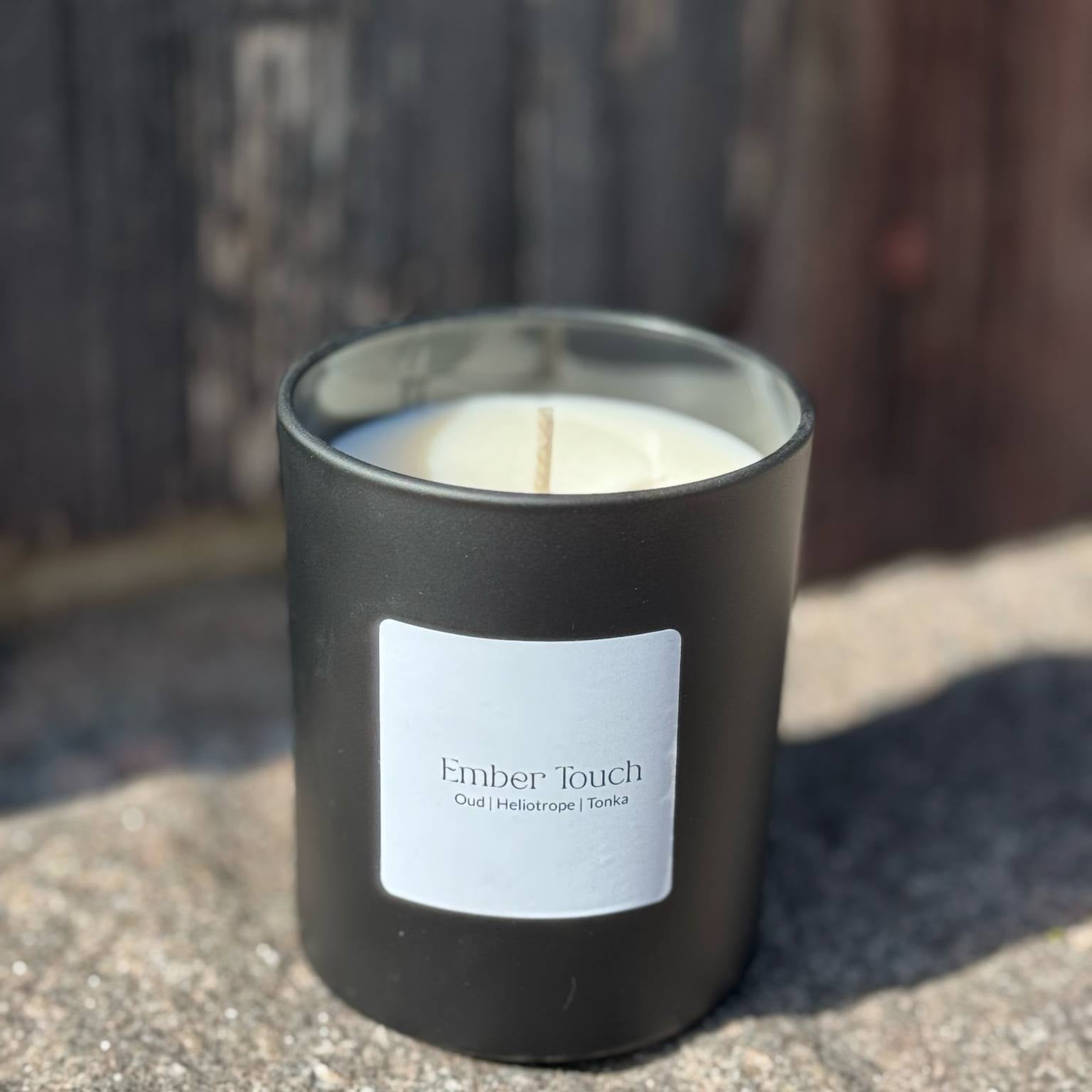 Doftljuset Ember Touch i mattsvart glas stående på solbelyst sten framför åldrat trä.