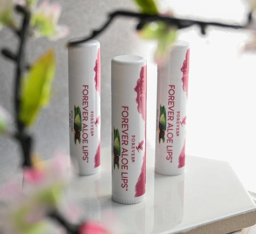 Forever Aloe Lips-produkter uppställda på vitt underlag med blommiga detaljer i förgrunden.