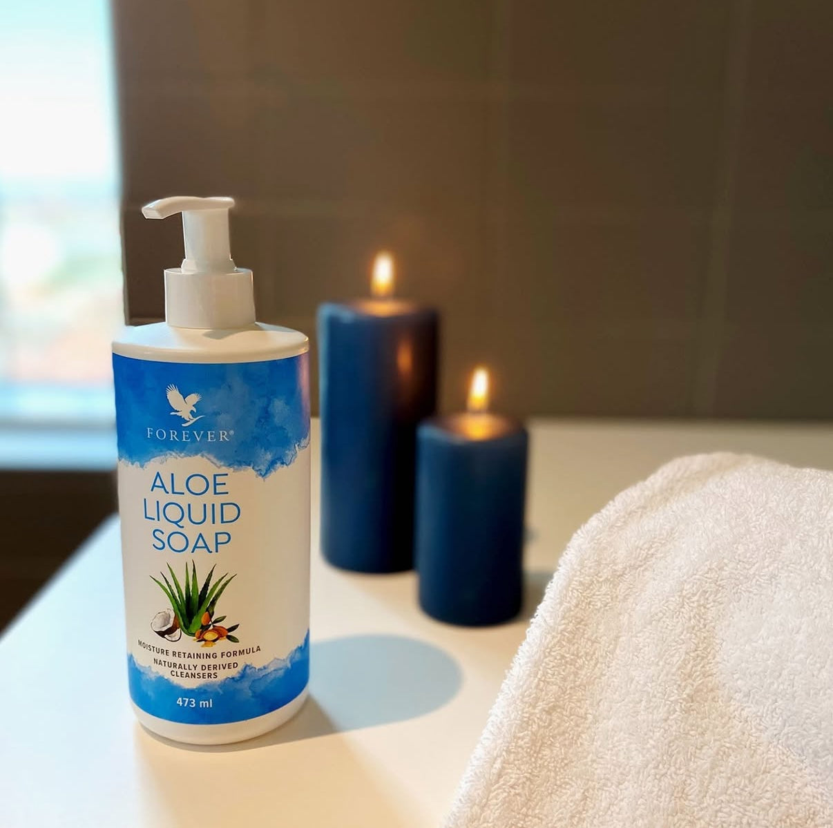 Forever Aloe Liquid Soap placerad på ett badrumsskåp tillsammans med tända ljus och handduk i en lugn vardagsmiljö.