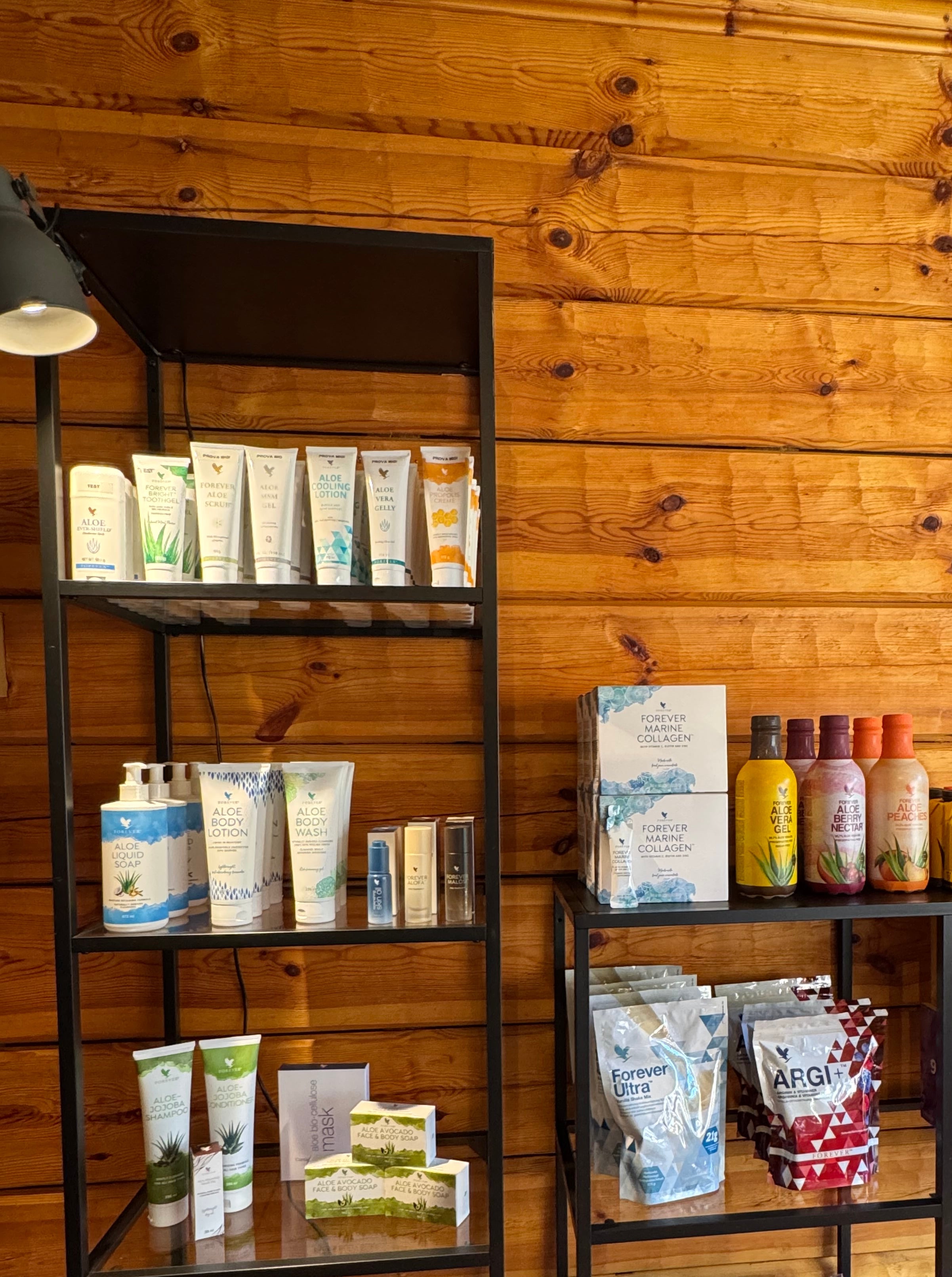 Forever Living-produkter uppställda på hyllor i gårdsbutiken, med aloe-drycker, hudvård och kosttillskott med en varm träinteriör bakom hyllorna.