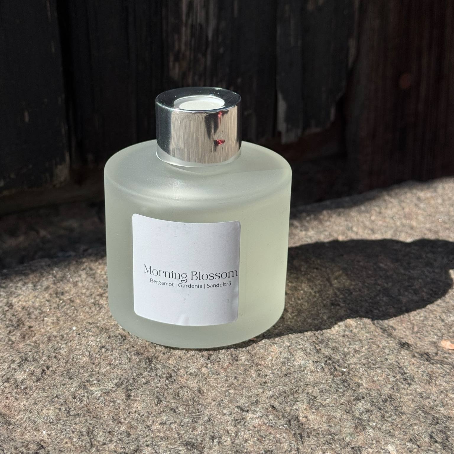 Morning Blossom doftpinnar i frostat glas med silvertopp framför sliten trävägg.